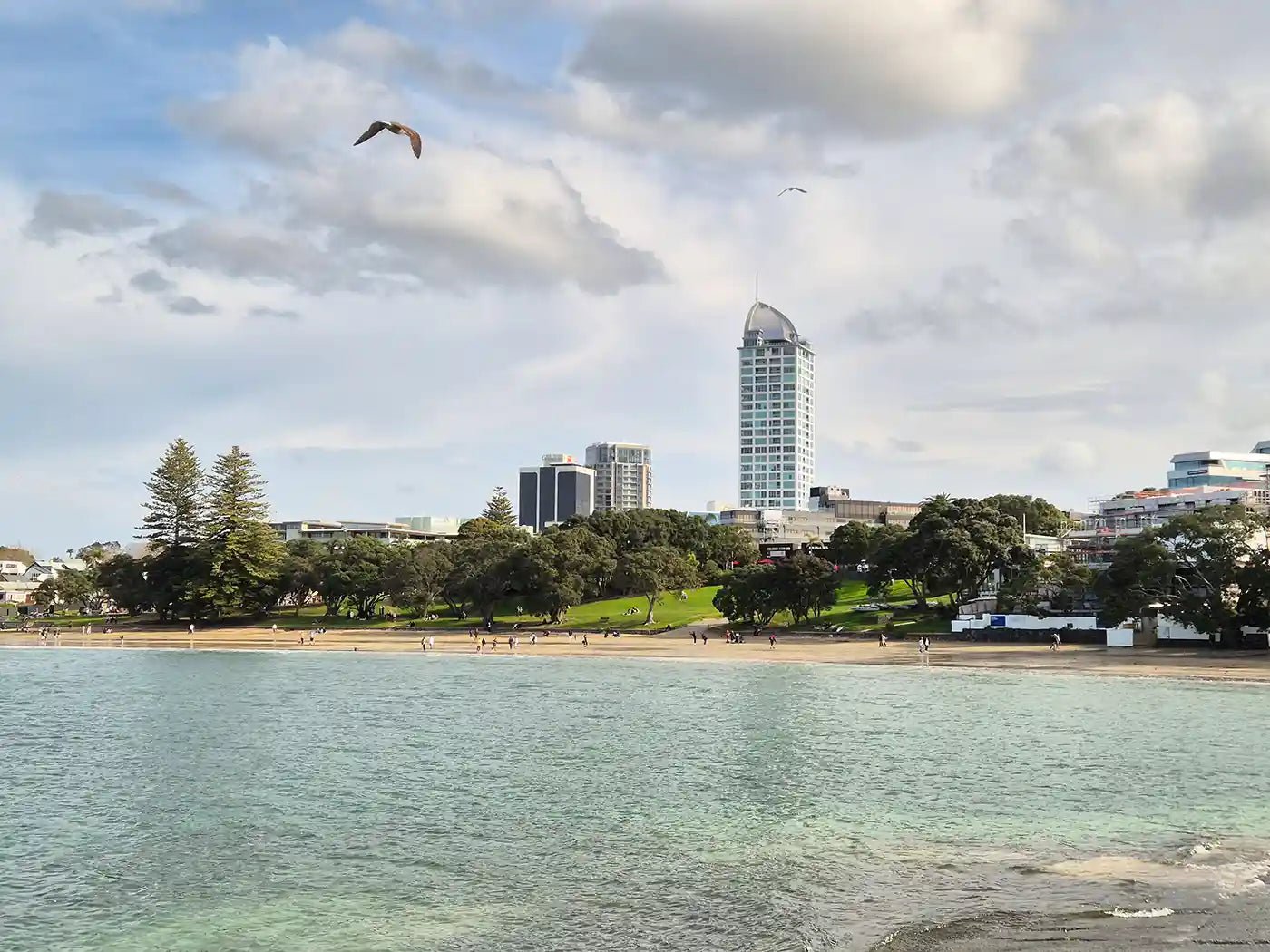 Image of Takapuna beach in Auckland, New Zealand. Three tall building in the background. Blue skies with mild cloud coverage.